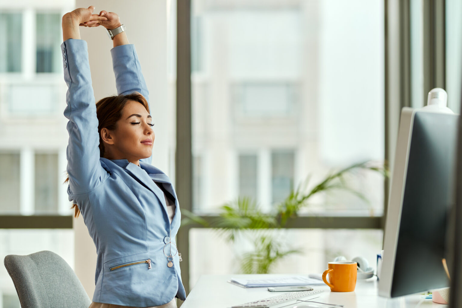 Young smiling businesswoman stretching with her eyes closed while working on a computer in the office.